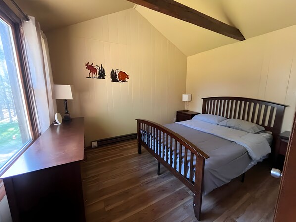 A bedroom with a wooden bed, a nightstand, a desk with a lamp, and a window.