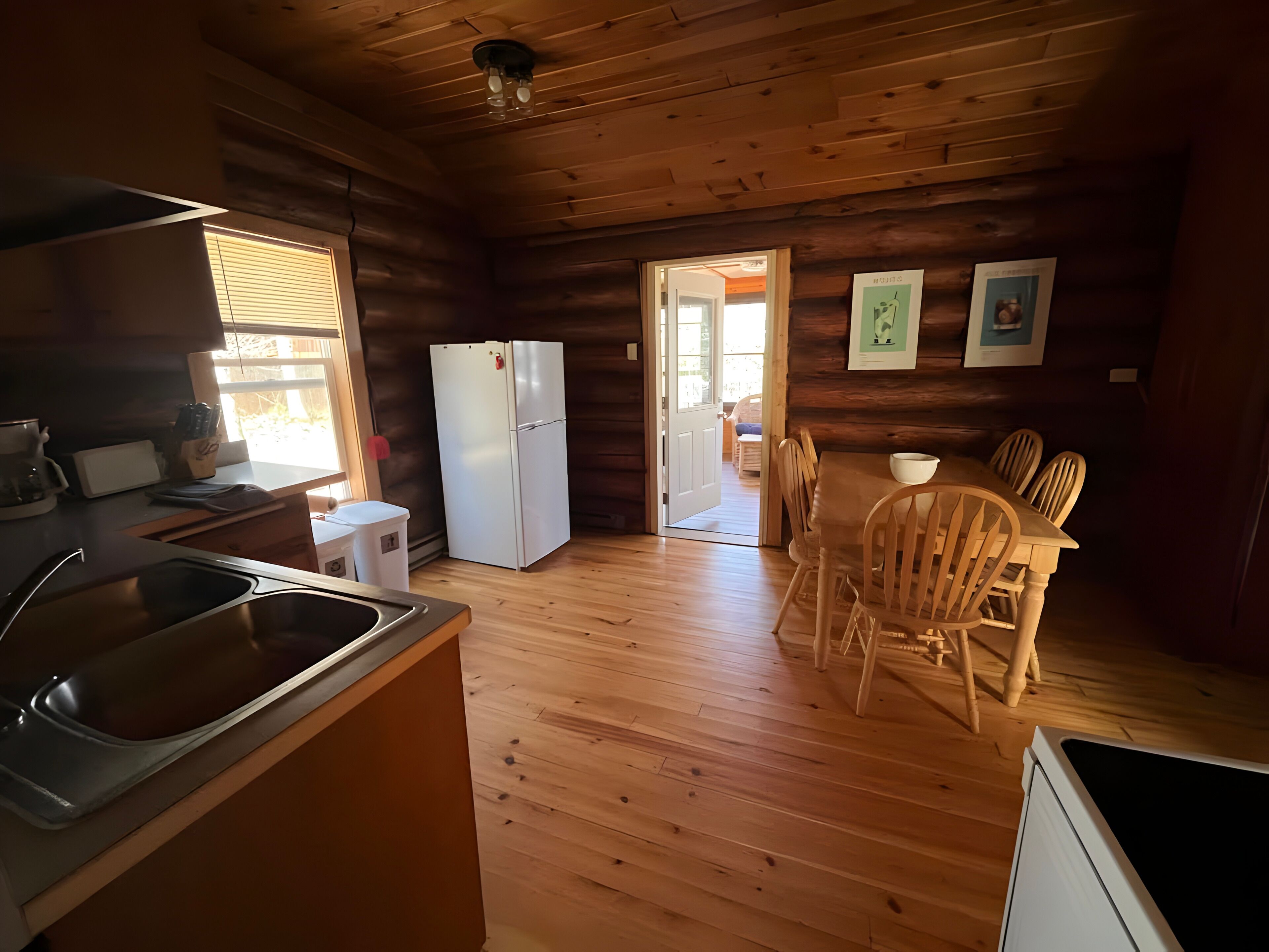 A wooden cabin interior with a kitchen, dining area, and a door leading outside.
