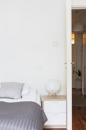 Un dormitorio con cama, mesita de noche con cajones, lámpara blanca y puerta que da a un baño.