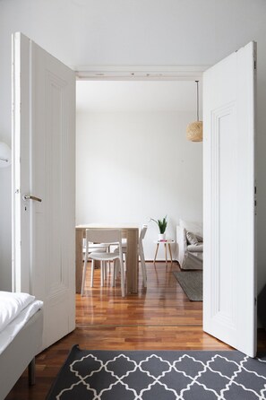 Une pièce comprenant un lit, un plancher en bois, une table à manger avec des chaises, une petite table avec une plante et un luminaire suspendu.