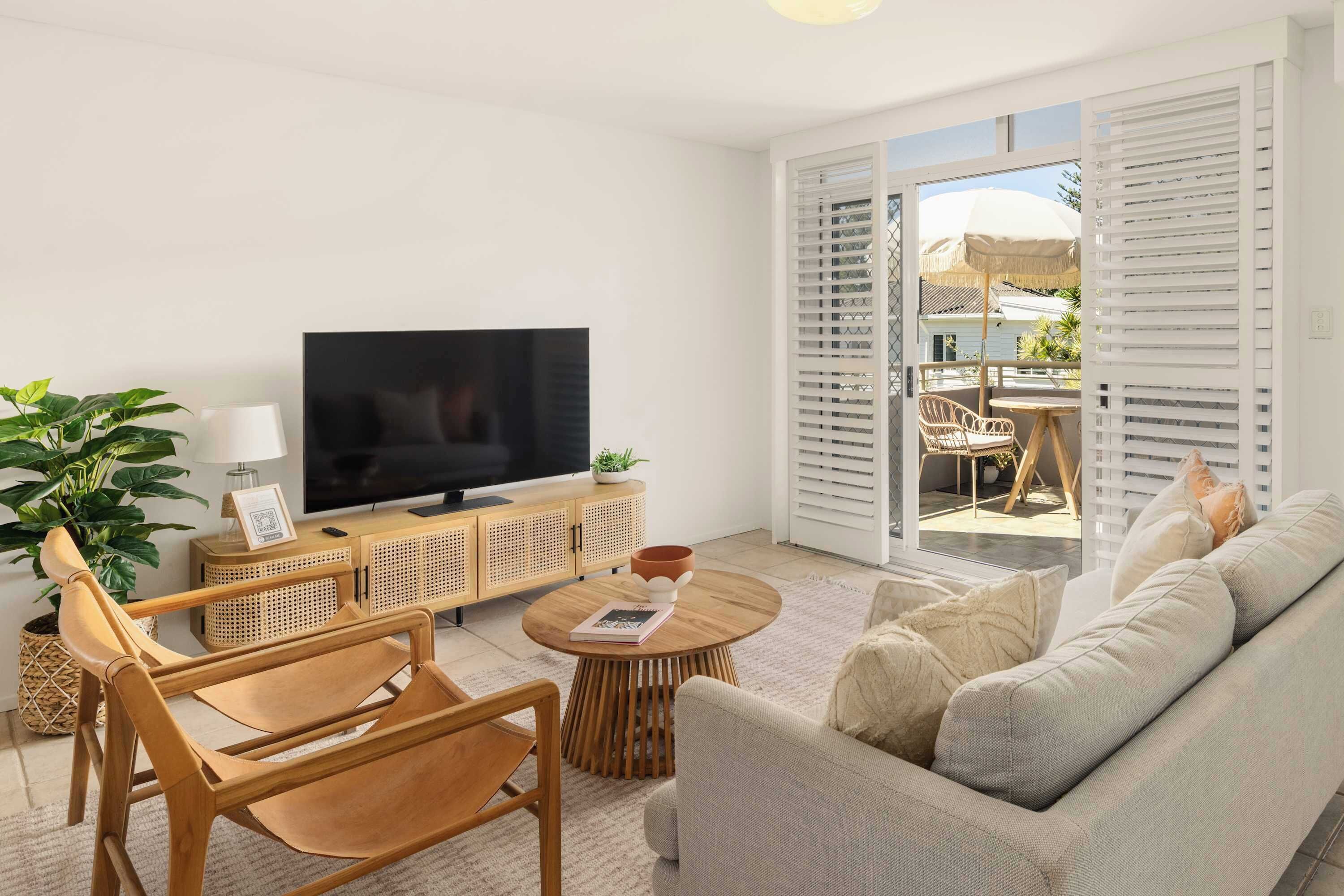 A living room with a flat-screen TV, wooden furniture, a sofa, and a view of an outdoor patio with a table and chairs.