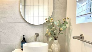 Een moderne badkamer met een ronde spiegel, een wit wastafeltje, een houten kastje en een vaas met bloemen.