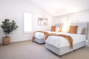 Une chambre avec deux lits, une plante en pot, un tableau encadré et une fenêtre avec des stores.