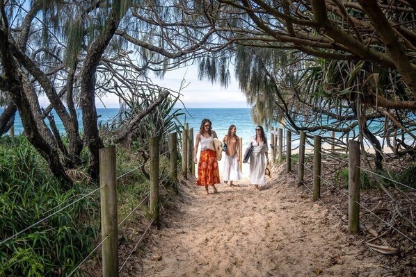 Un sentier sablonneux menant à une plage, bordé d’arbres et entouré d’une clôture en bois.
