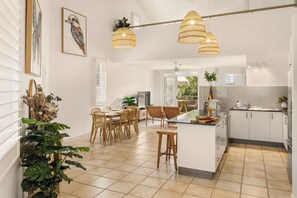A modern kitchen with white cabinets, a central island, and a dining area with wicker chairs. - Spacious Seaside Oasis on Dicky Beach (Dicky Beach)