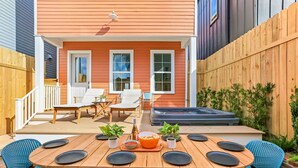 Une terrasse avec une table en bois dressée pour les repas, des chaises d’extérieur, un jacuzzi et un petit coin jardin.
