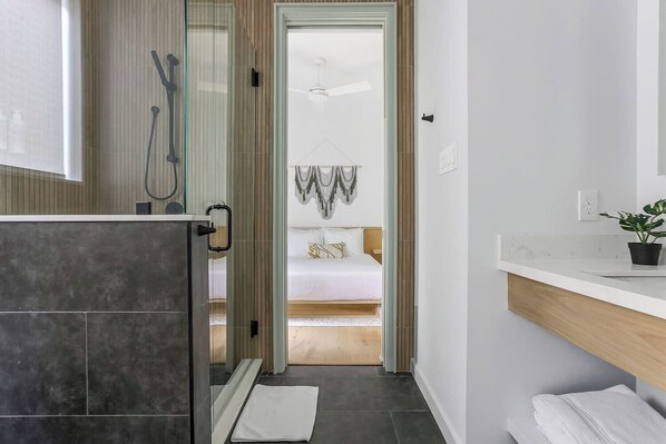 Un baño moderno con ducha, inodoro y lavamanos. Se ve una cama a través de una puerta de vidrio que da a un dormitorio.