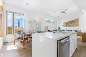 A modern kitchen with a large island, wooden chairs, pendant lights, and a view of the outdoors.