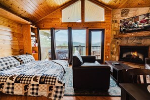 A cozy cabin interior with a bed, seating area, fireplace, and large windows.