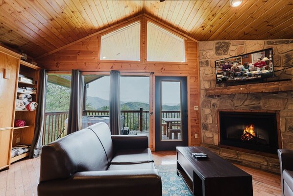 Interior acogedor de una cabaña con chimenea, un sofá de cuero, televisor y un balcón con vistas a las montañas.