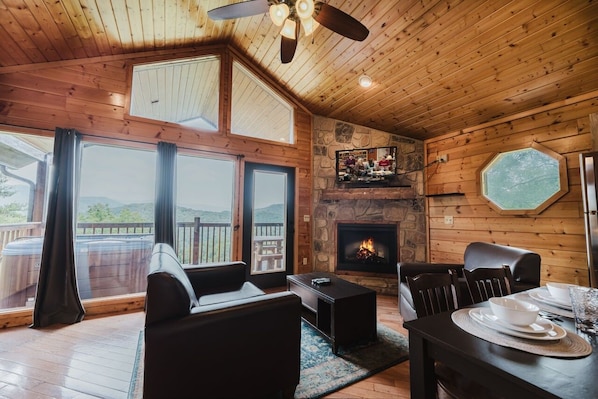 Interior acogedor de una cabaña con chimenea, paredes de madera, un comedor y ventanas grandes con vistas a una terraza.