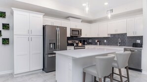 Uma cozinha moderna com armários brancos, eletrodomésticos de aço inoxidável, uma ilha central com banquetas e um backsplash de azulejos pretos.