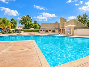 A swimming pool with lounge chairs and umbrellas, surrounded by a building with a flat roof and a fence.