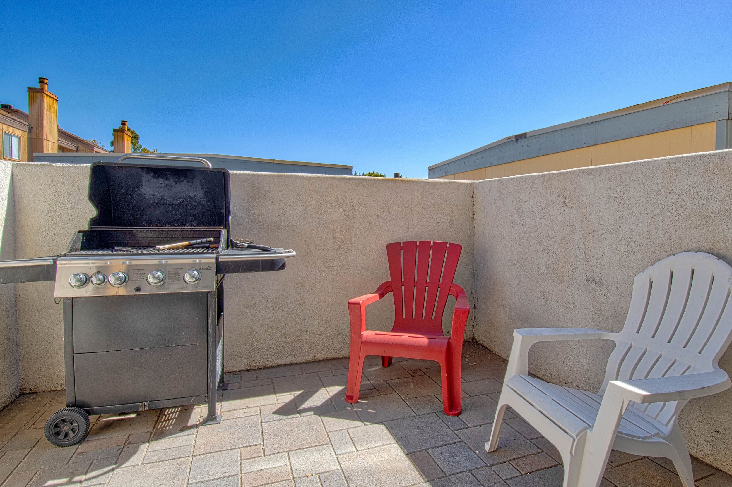 Un patio con una parrilla, dos sillas Adirondack y piso de baldosas.