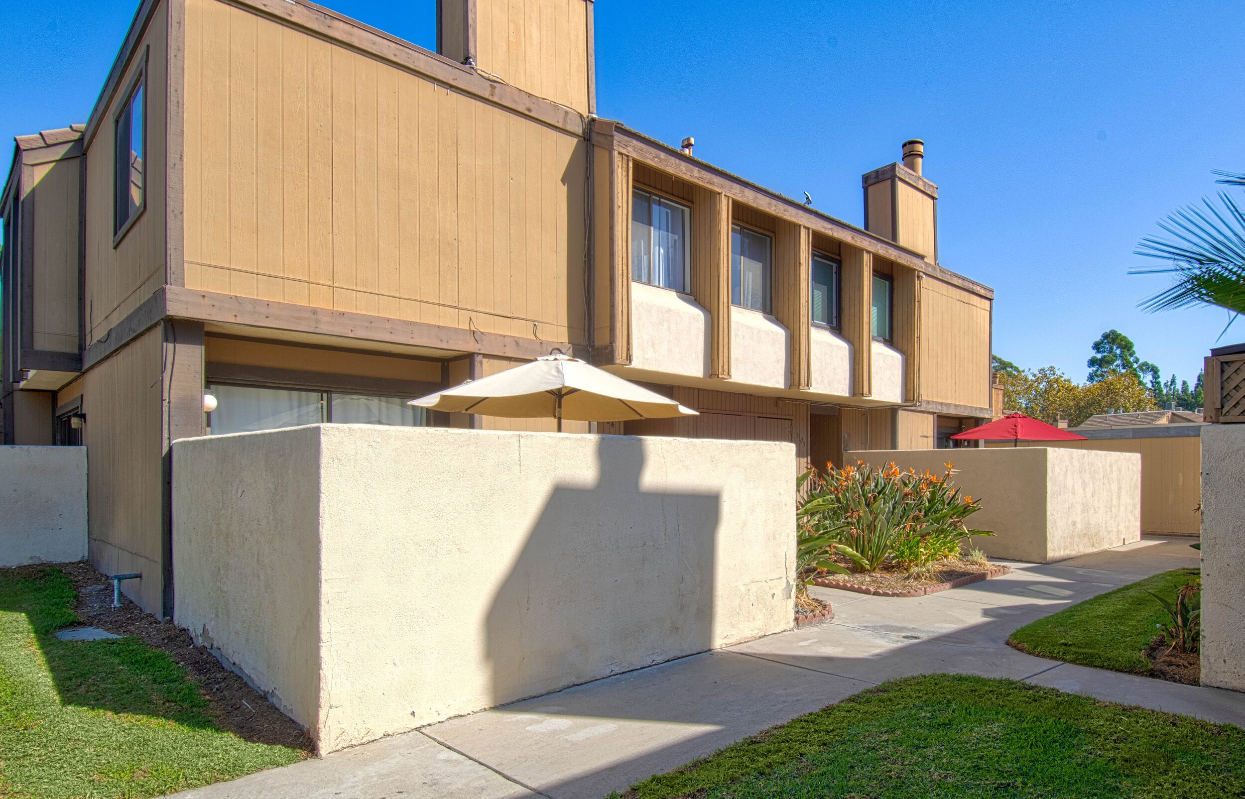 Un bâtiment moderne doté d’un toit plat, de grandes fenêtres et d’un chemin en béton menant à un patio couvert agrémenté d’un parasol rouge.