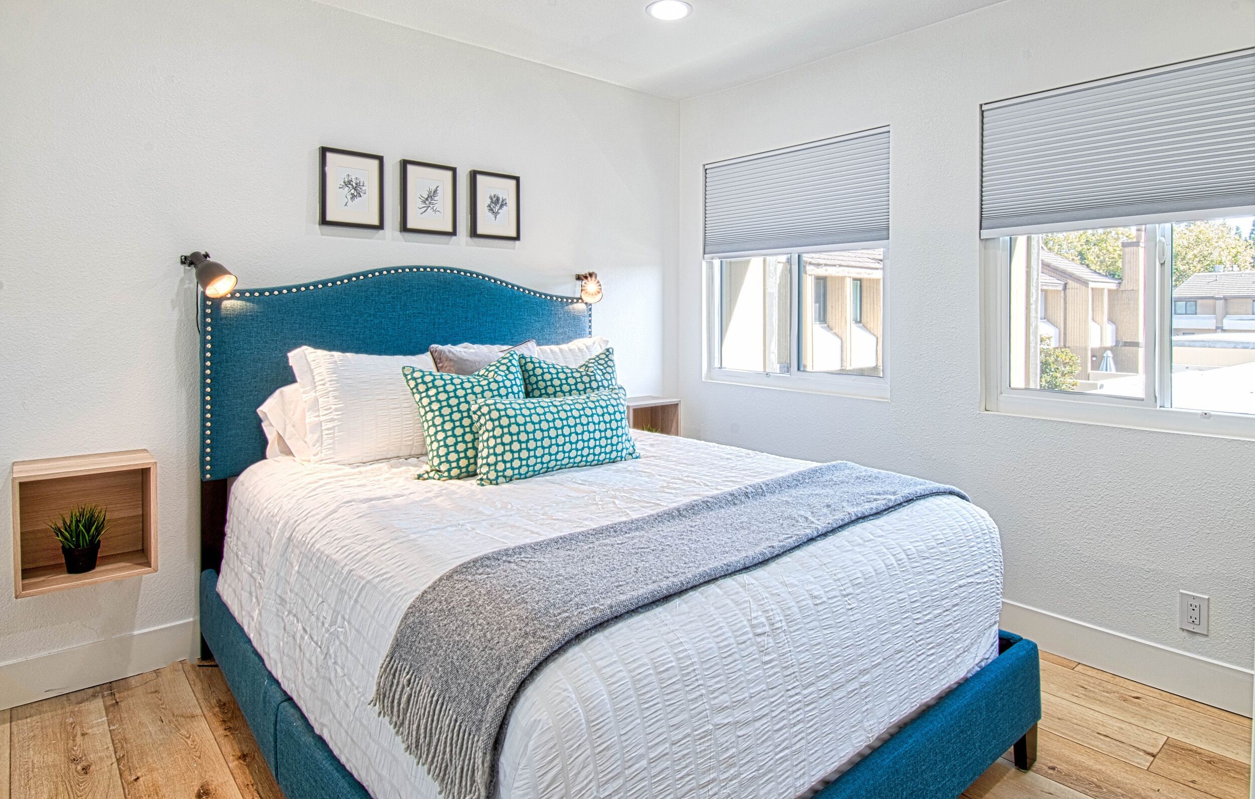A bedroom with a large bed, a headboard with lights, a window with blinds, and a small plant on a shelf.