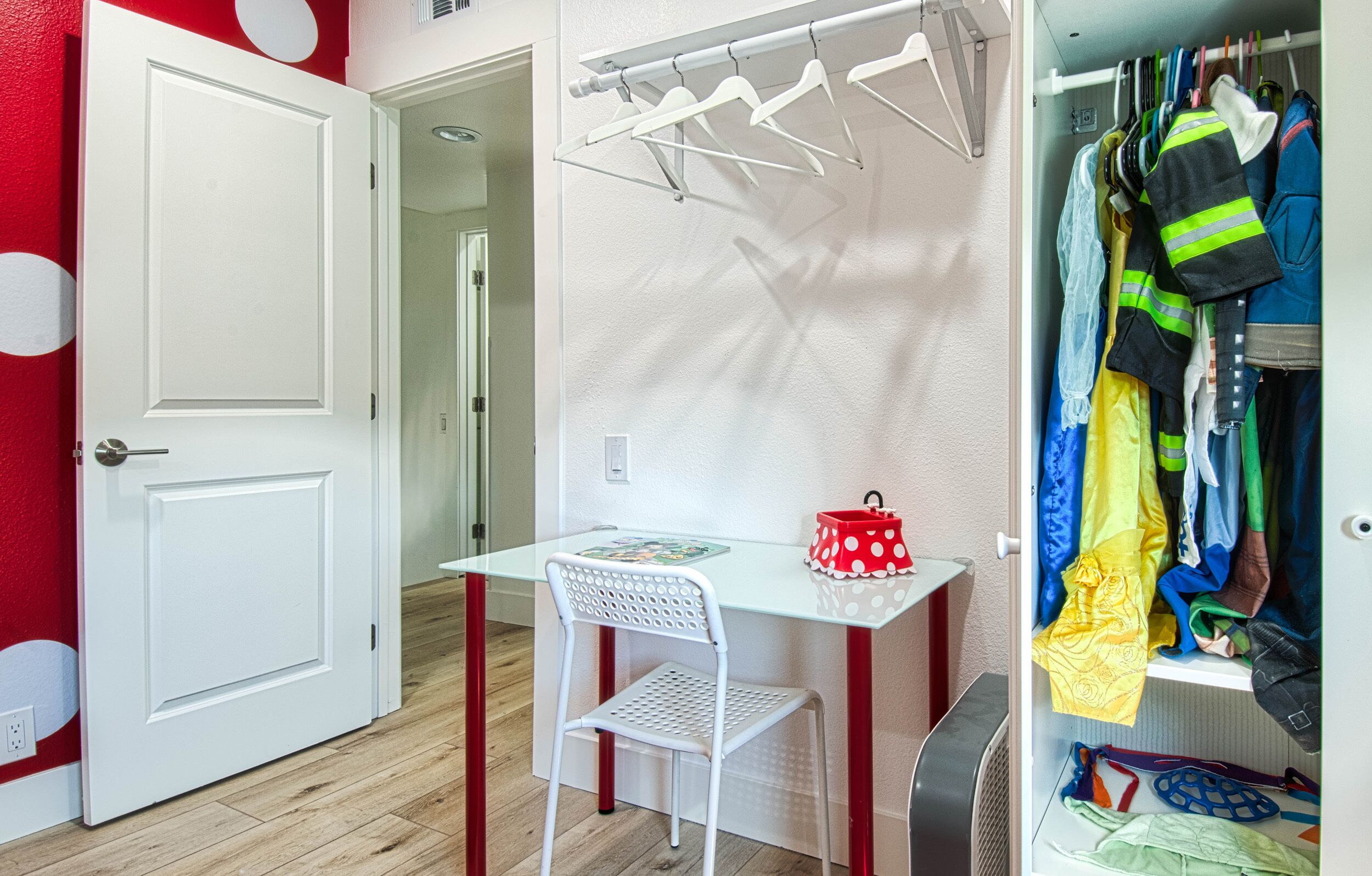 Une pièce avec une porte blanche, une petite table avec une chaise, un sac rouge à pois et un portant à vêtements sur lequel sont suspendus différents articles.