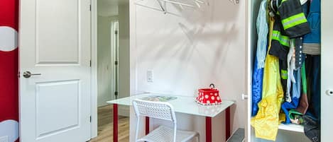 Une pièce avec une porte blanche, une petite table avec une chaise, un sac rouge à pois et un portant à vêtements sur lequel sont suspendus différents articles.
