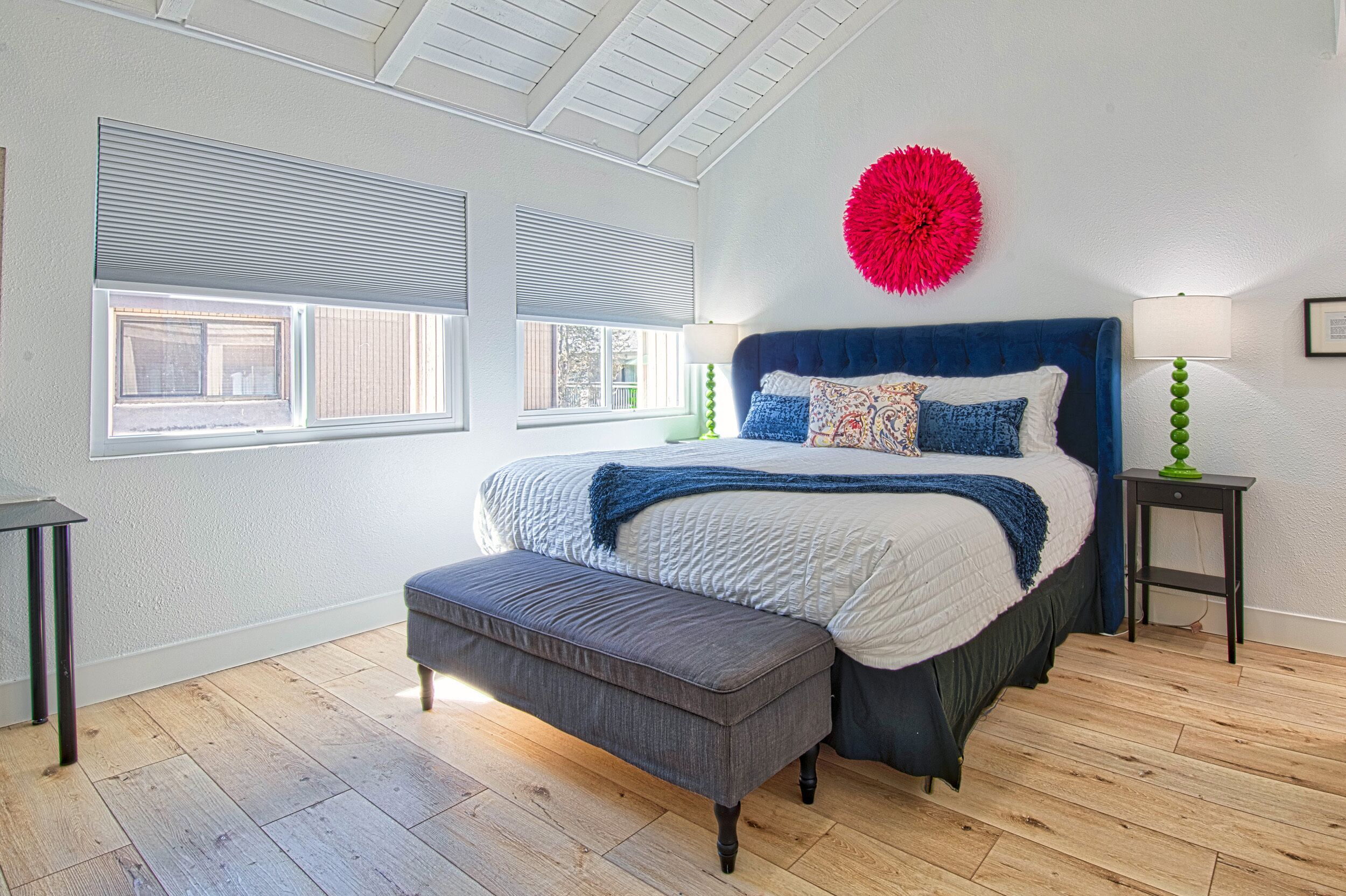 Un dormitorio con una cama grande, un banco, dos ventanas, un adorno rojo y una lámpara verde.