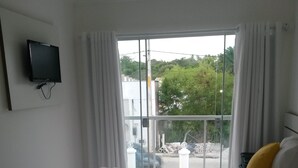 A hotel room with a large bed, white curtains, a flat-screen TV mounted on the wall, and a view of the outdoors through a large window. - Porto Seguro-Condomínio Mont Carmelo Ap 5A (Porto Seguro)