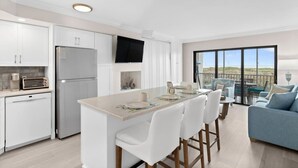 A modern kitchen with white cabinets, a built-in refrigerator, a flat-screen TV, and a dining area with white chairs and a light-colored table.