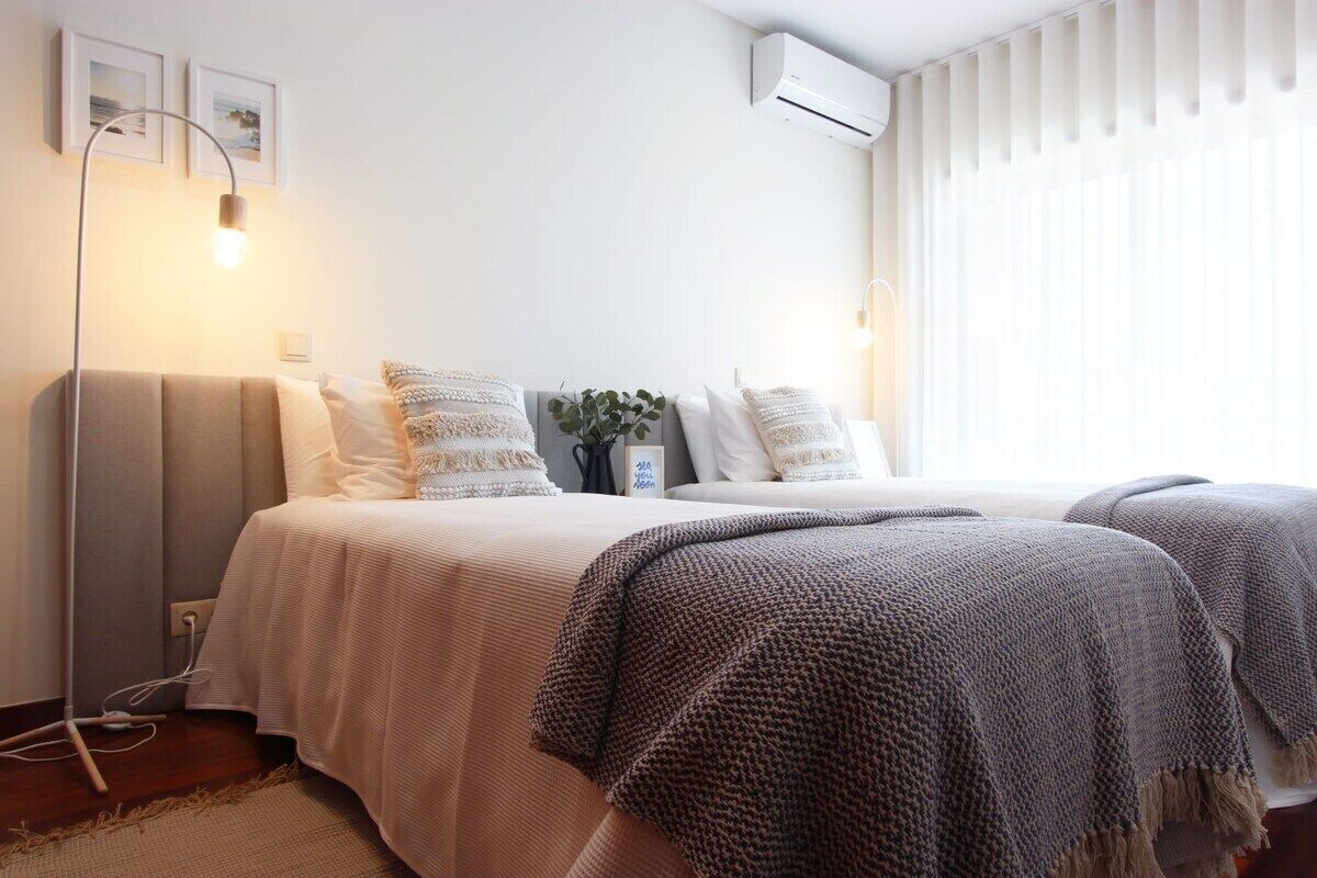 Quarto com duas camas, luminária de cabeceira, ar condicionado fixado na parede e janela com persianas.