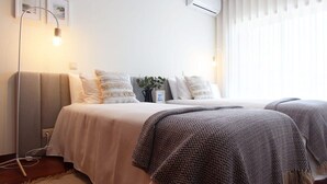 A bedroom with two beds, a bedside lamp, a wall-mounted air conditioner, and a window with blinds.