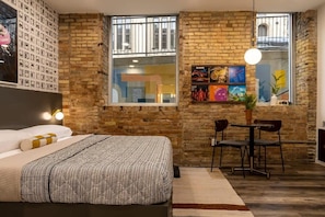 A bedroom with a bed, a small table with chairs, a window with artwork, and a brick wall.