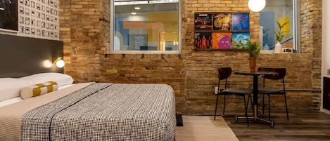 A bedroom with a bed, a small table with chairs, a window with artwork, and a brick wall.