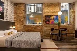 Une chambre avec un lit, une petite table avec des chaises, une fenêtre décorée d’œuvres d’art et un mur en briques.