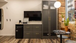 Una habitación moderna con televisión de pantalla plana, refrigerador compacto, cafetera, mesa de comedor con sillas y un amplio ventanal con vista a un edificio.
