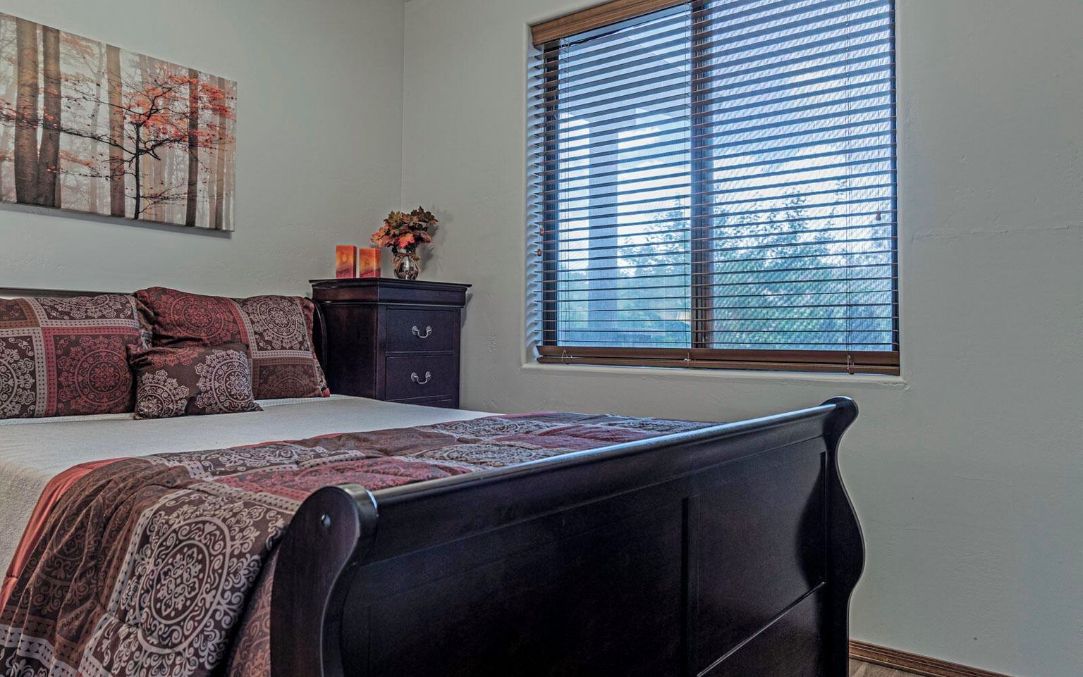 Une chambre à coucher avec un lit, une commode, une fenêtre avec des stores et un tableau accroché au mur.