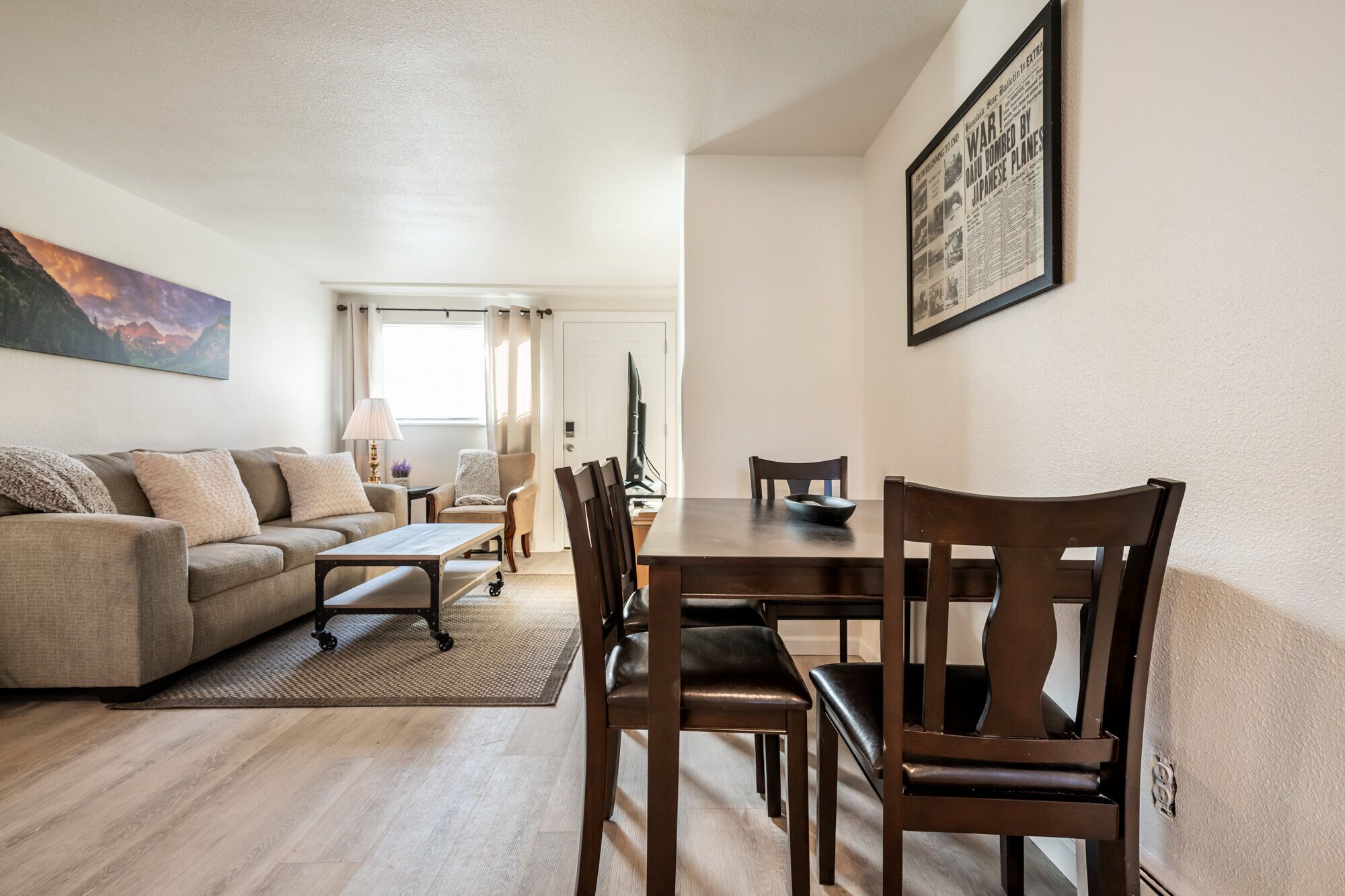 A living room with a sofa, coffee table, and dining table with chairs. There is a painting on the wall and a framed newspaper on the wall.