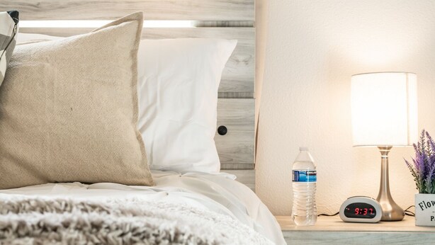 Ein ordentlich gemachtes Bett mit weißer Bettwäsche und einer beigen Tagesdecke, eine Nachttischlampe, eine Wasserflasche und eine digitale Uhr, die 9:37 Uhr anzeigt.