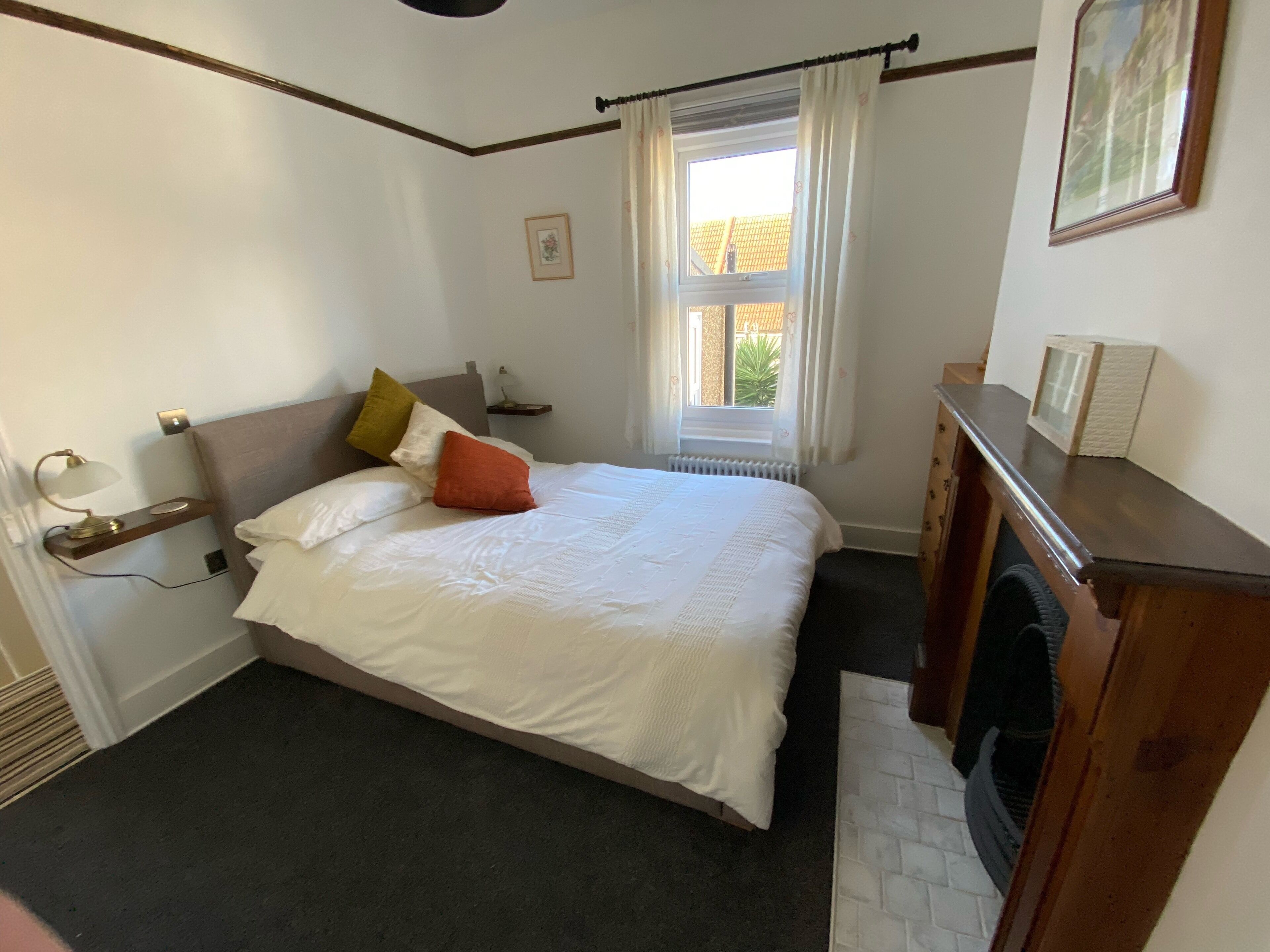 A neatly made bed with white linens and multiple pillows, a bedside table with a lamp, a small shelf, and a window with curtains.