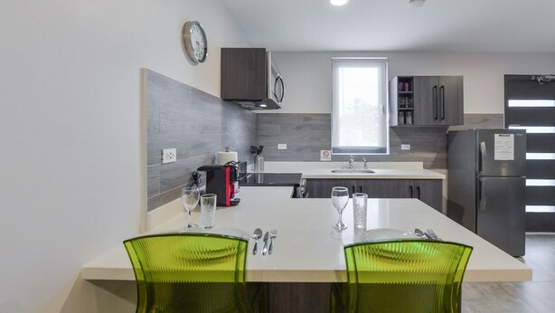 A modern kitchen with a central island, two green chairs, a coffee machine, and a refrigerator.