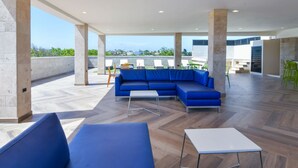 A spacious outdoor seating area with blue sofas and a white table, surrounded by pillars and a view of the cityscape.