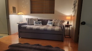 A cozy bedroom with a grey sofa bed, patterned pillows, a small bedside table with a lamp, and a window with blinds.