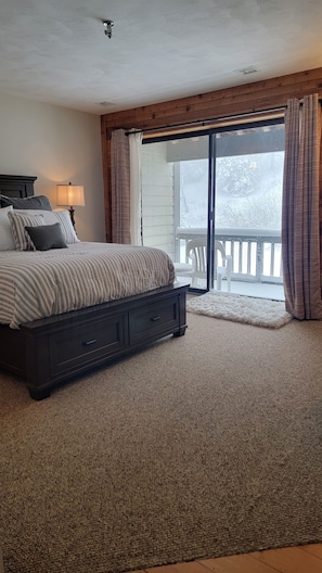 A bedroom with a large bed, a nightstand, a lamp, and a view of a snowy outdoor area through a sliding glass door.