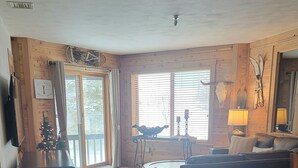 A spacious living room with a wooden floor, a sofa, a coffee table, and a dining table. There is a small Christmas tree and a sign that reads "Ski Lodge."