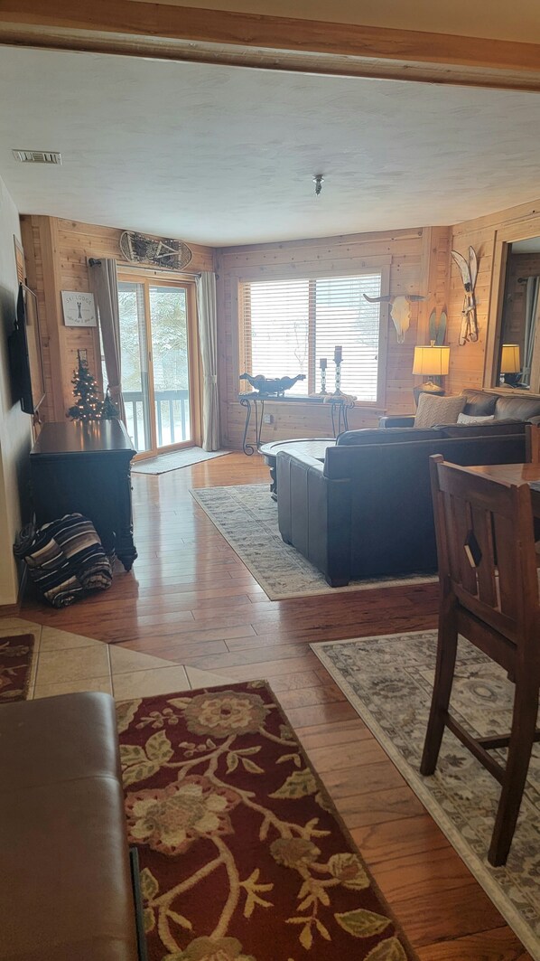 A spacious living room with a wooden floor, a sofa, a coffee table, and a dining table. There is a small Christmas tree and a sign that reads "Ski Lodge."