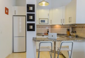 A compact kitchen with white cabinets, a microwave, a refrigerator, a sink, and a laptop on the counter. There are two bar stools and a wine bottle.
