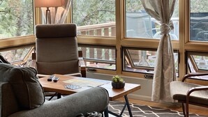 Een kamer met een groot raam, een stoel, een tafel met een lamp en uitzicht op een bergachtig landschap.