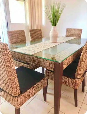 Un coin repas avec une table à plateau en verre, entourée de chaises en osier dotées de coussins noirs.