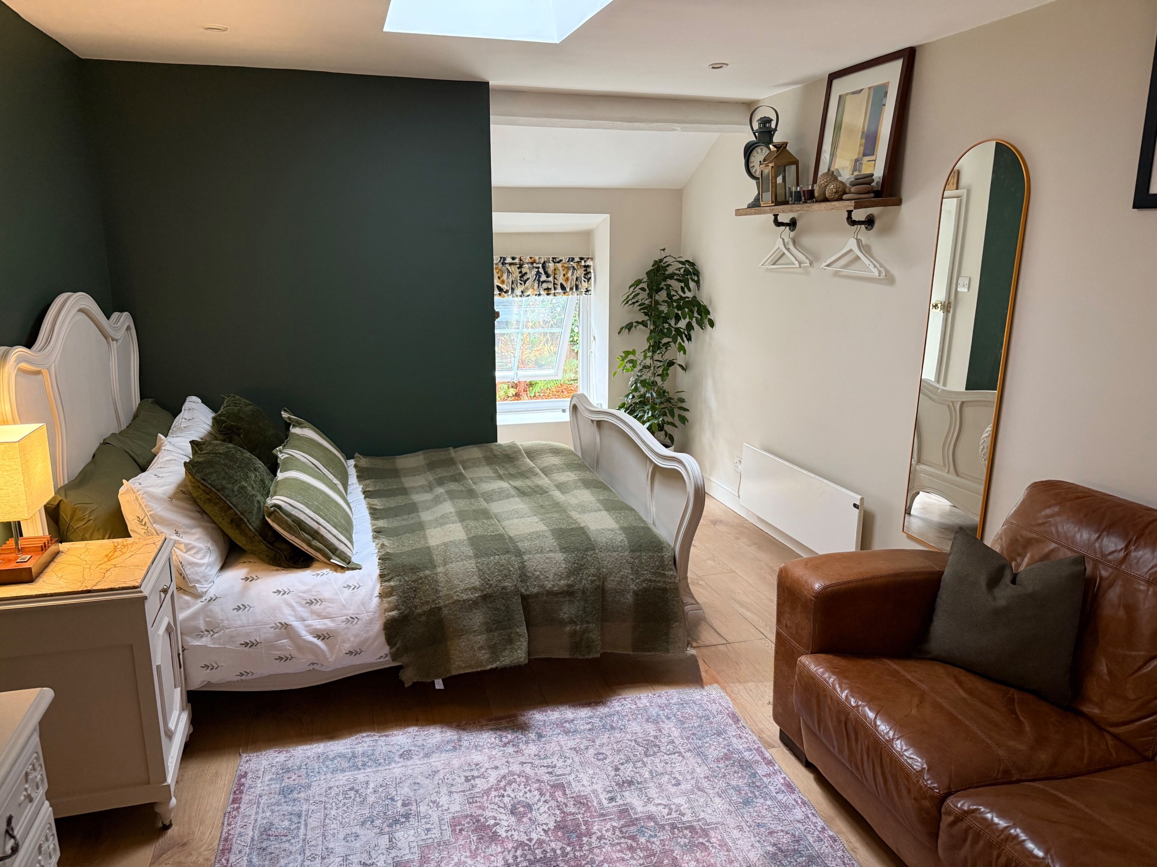 A bedroom with a bed, a dresser, a brown leather couch, a large mirror, and a window with a view of greenery.