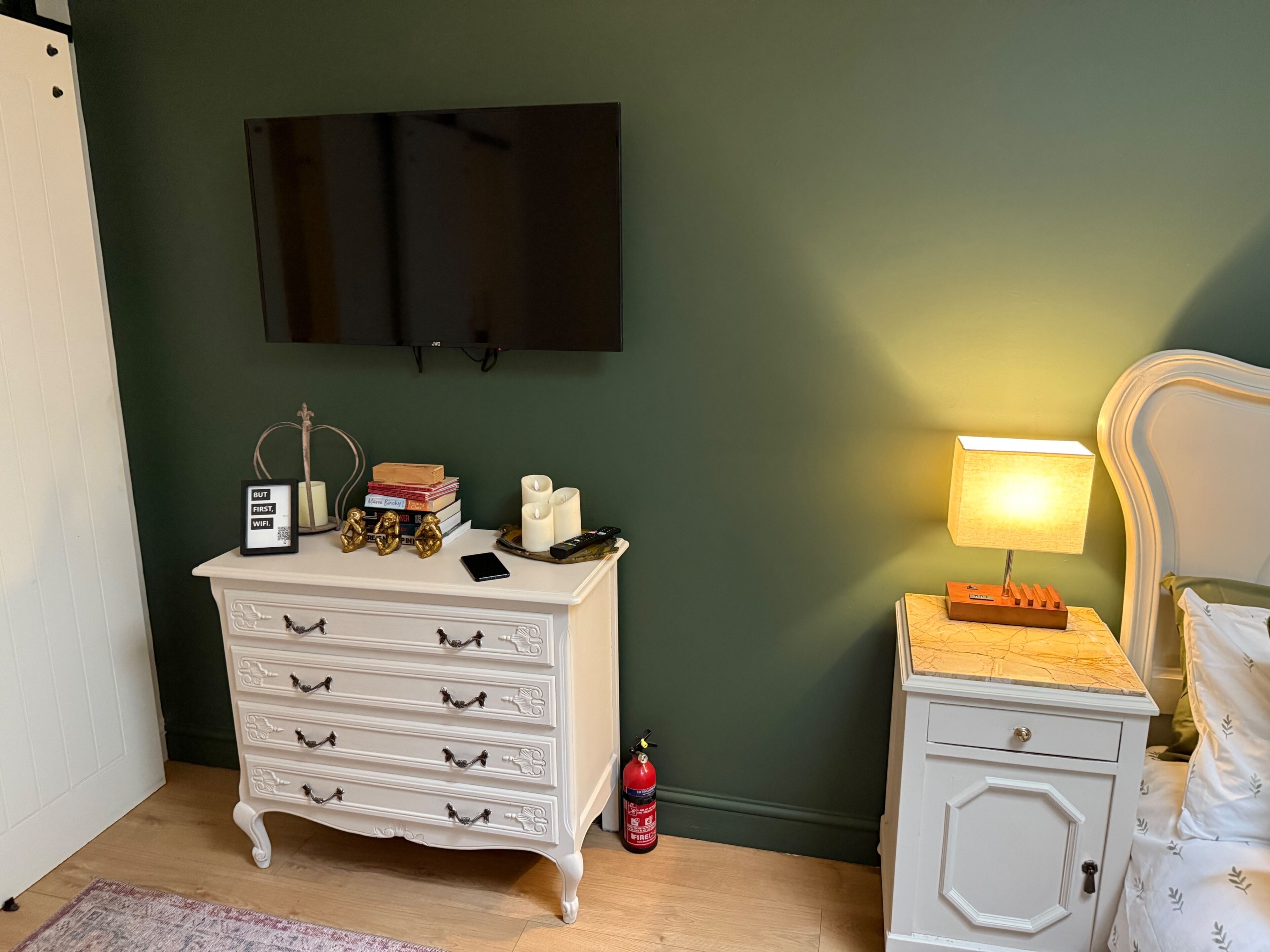 A bedroom with a white dresser, a bedside table with a lamp, a TV mounted on the wall, and a fire extinguisher.