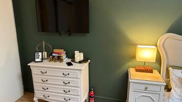 A bedroom with a white dresser, a bedside table with a lamp, a TV mounted on the wall, and a fire extinguisher.