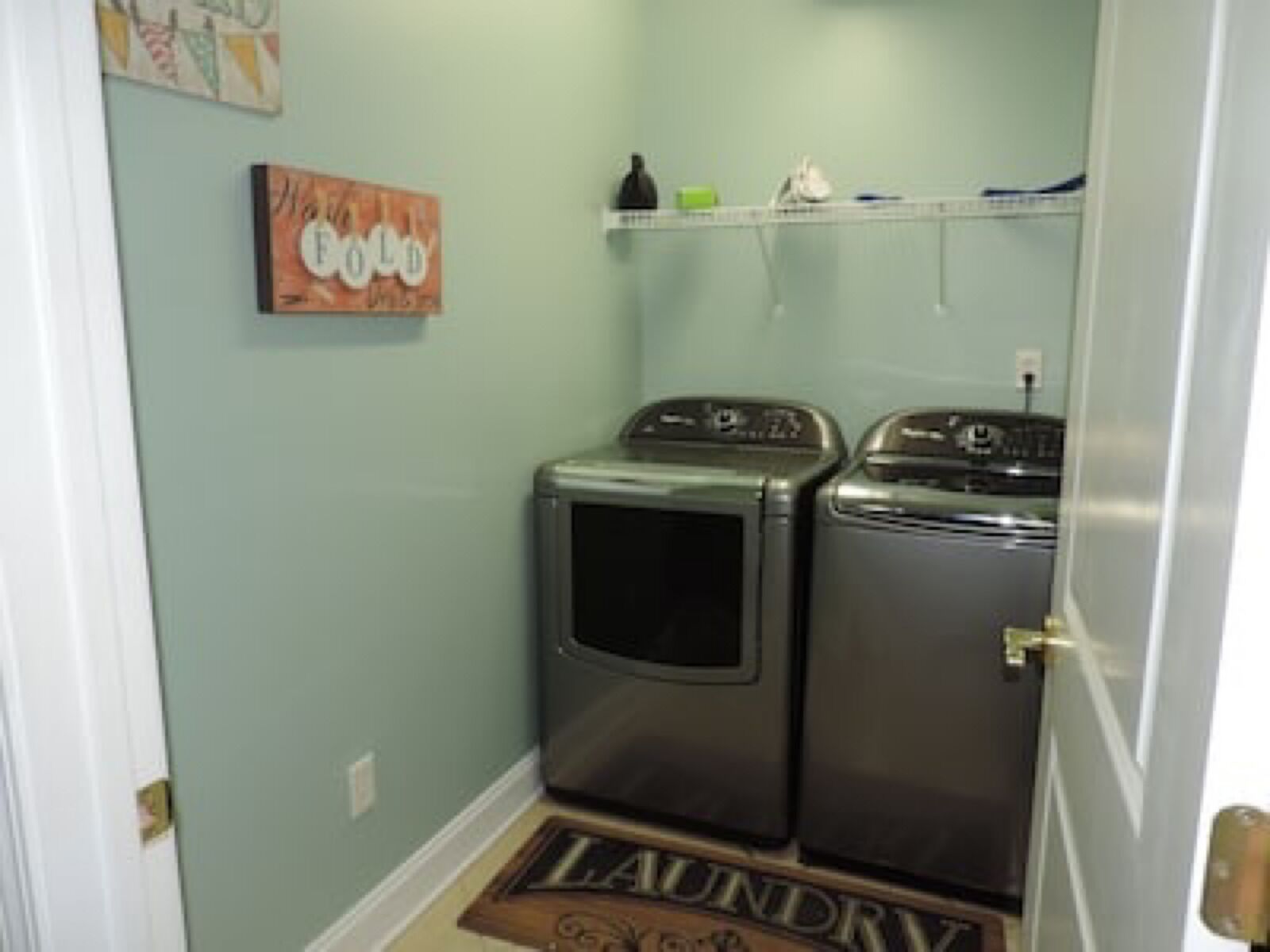 A laundry room with two stacked washers and dryers, a shelf with laundry supplies, and a 'Laundry' mat on the floor.