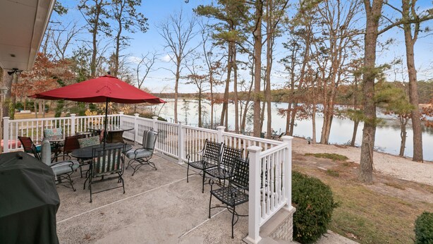 A covered patio with outdoor furniture and a red umbrella, overlooking a lake with trees and a white fence.