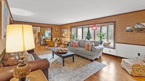 A spacious living room with a grey sofa, a coffee table, and a lamp. There is a dining area in the background with a table and chairs.